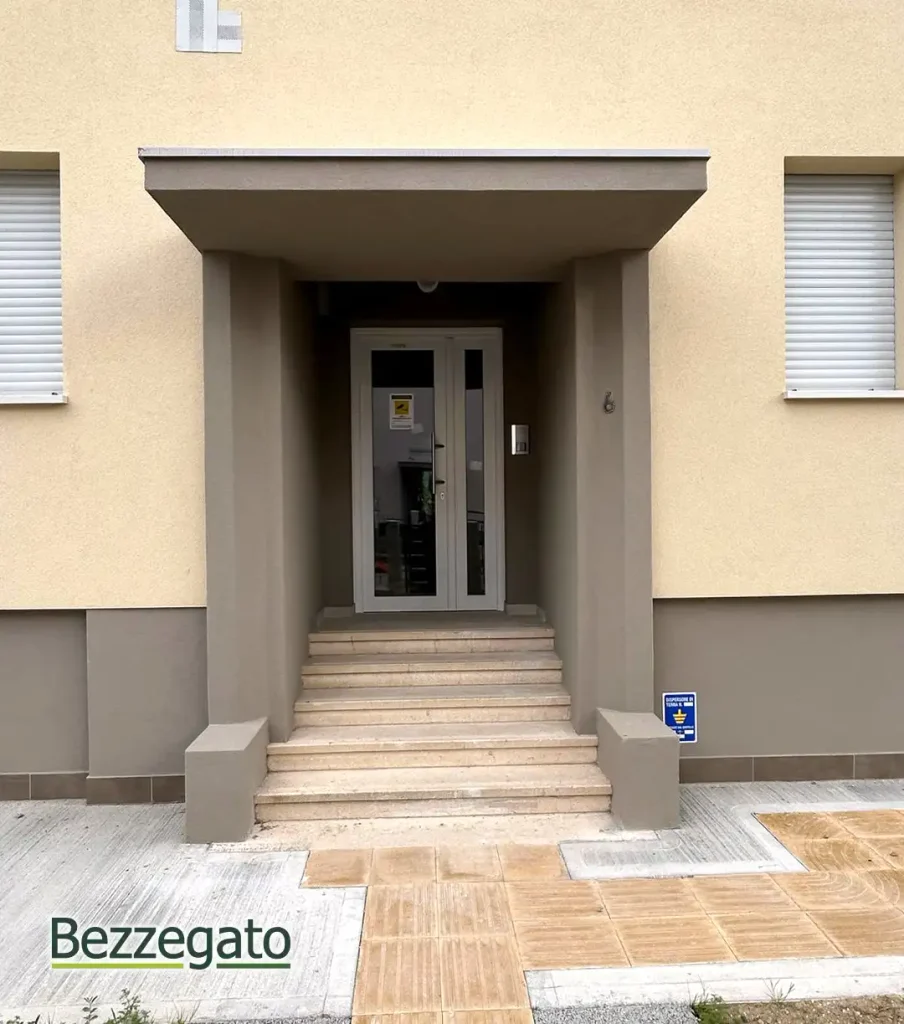 INGRESSO copia_web | Bezzegato Entrance to a beige building with a small covered porch and a set of stone stairs leading to glass double doors (security/intercom on the right).