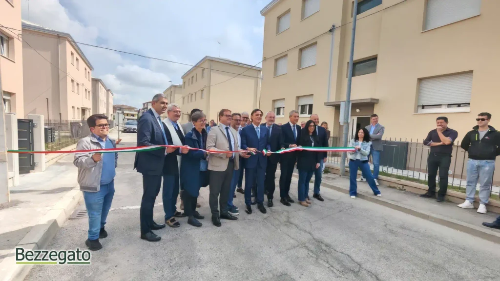 WhatsApp Image 2026-05-08 at 151842_upscayl_2x_high-fidelity-4x_web | Bezzegato Group of formally dressed people cutting a red, white, and green ribbon outside beige apartment buildings during a ribbon-cutting ceremony on a sunny day.
