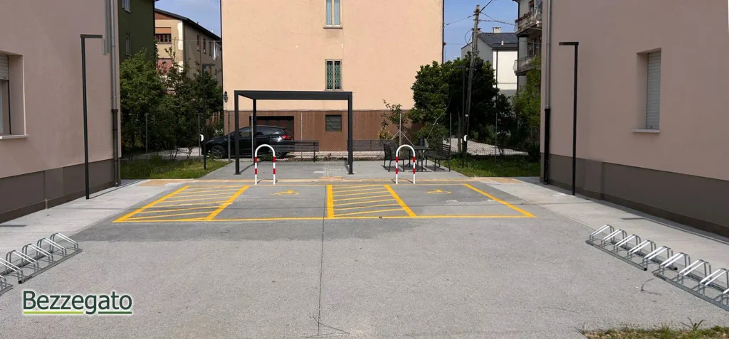 WhatsApp Image 2026-05-08 at 155255 copia_web | Bezzegato Parking lot with two yellow-striped accessible parking spaces, white-and-red wheelchair barriers, a metal shelter, benches, and bike racks, set against beige residential buildings in the background.