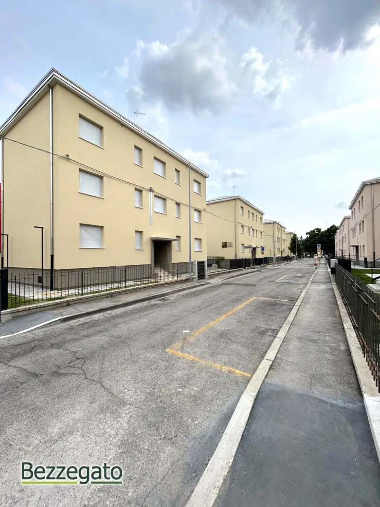 WhatsApp Image 2026-05-08 at 155507 copia_web | Bezzegato Residential street with beige apartment buildings, metal railings, and a paved road; minimal traffic and clear sky.