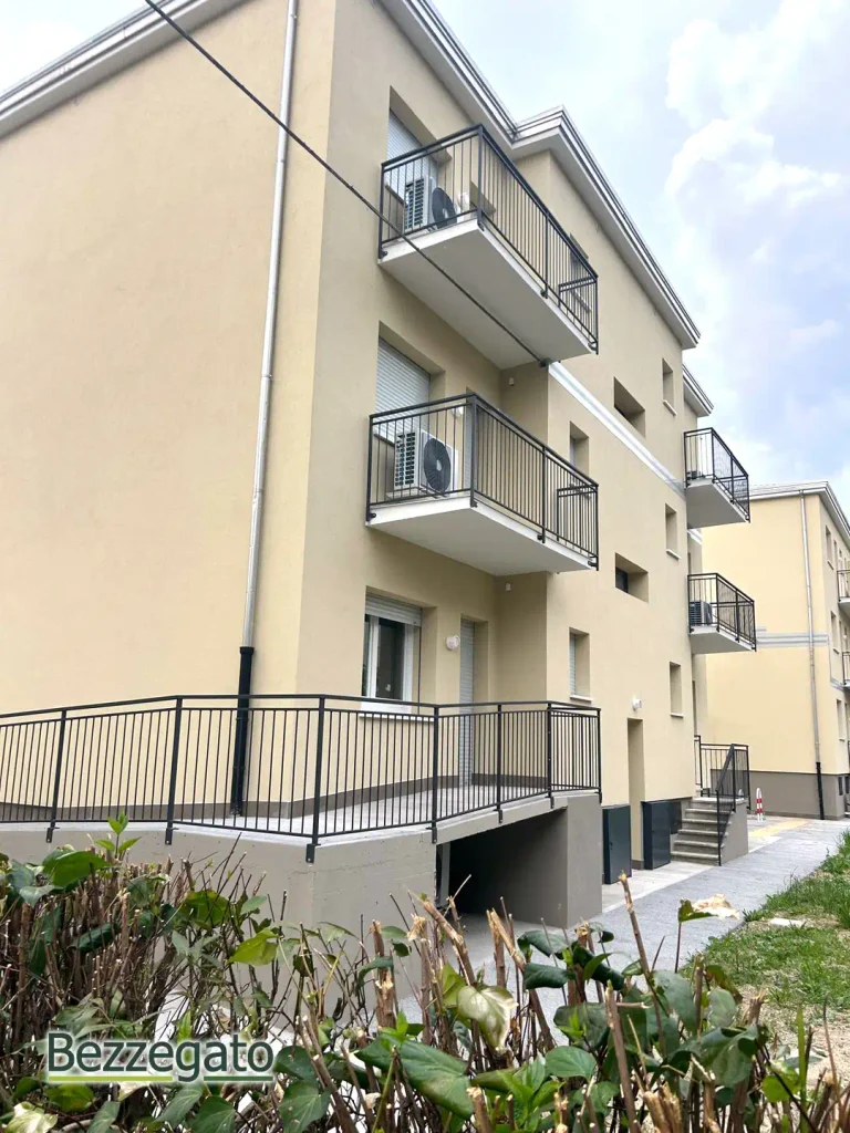dettagli esterni copia_web | Bezzegato Exterior view of a beige multi-story apartment building with staggered balconies and black railings, air conditioners on each balcony, against a cloudy sky, Bezzegato watermark bottom left.