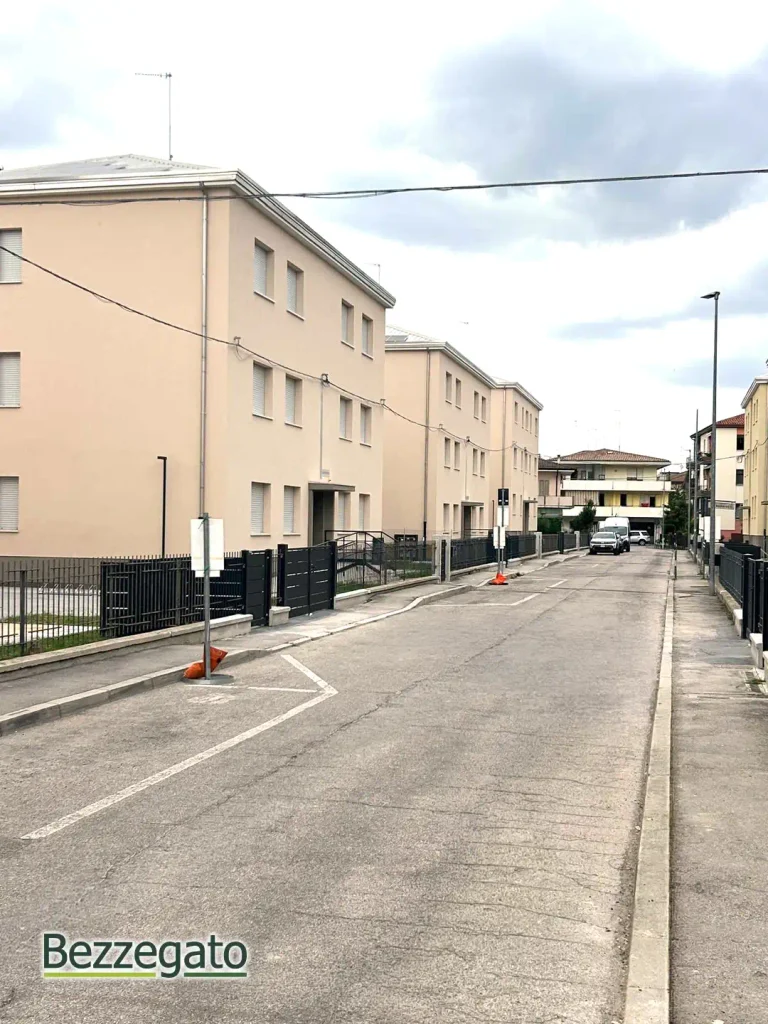 palazzine copia_web | Bezzegato Residential street with beige multi‑story apartment buildings, fenced yards, and a parked car in the distance under a cloudy sky.