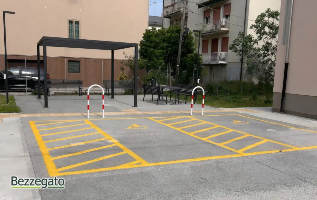 parcheggi copia_upscayl_2x_high-fidelity-4x_web | Bezzegato Parking lot with two handicapped spaces marked in yellow stripes, white safety barriers, and a seating area under a black metal pergola near residential buildings.