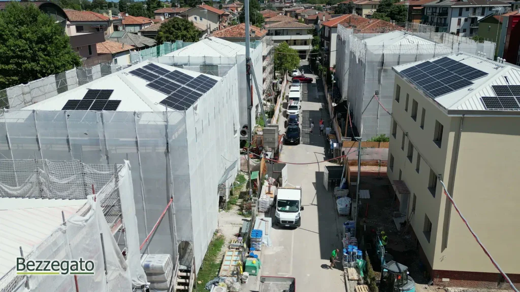 vlcsnap-2026-10-28-18h41m32s880_web | Bezzegato Aerial view of a narrow street between buildings under construction, with white scaffolding and solar panels on the rooftops, construction materials along the sidewalk, and parked cars below.