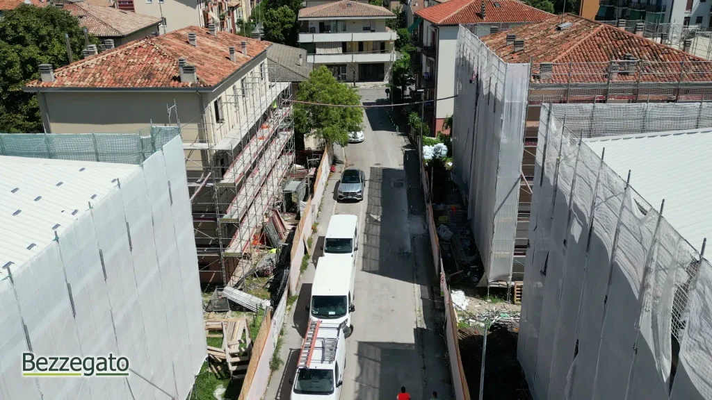 vlcsnap-2026-10-28-18h41m49s670_web | Bezzegato Aerial street view of a construction site flanked by buildings with scaffolding, netting, and white vans parked along the road.
