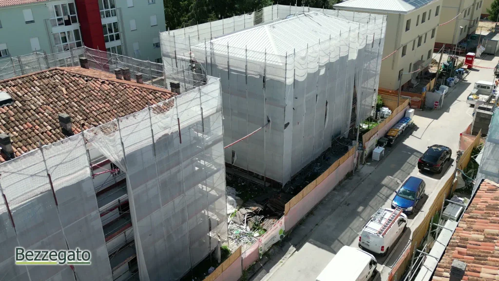 vlcsnap-2026-10-28-18h42m18s078_web | Bezzegato Aerial view of a construction site with netted scaffolding around a building beside a narrow street with parked cars.
