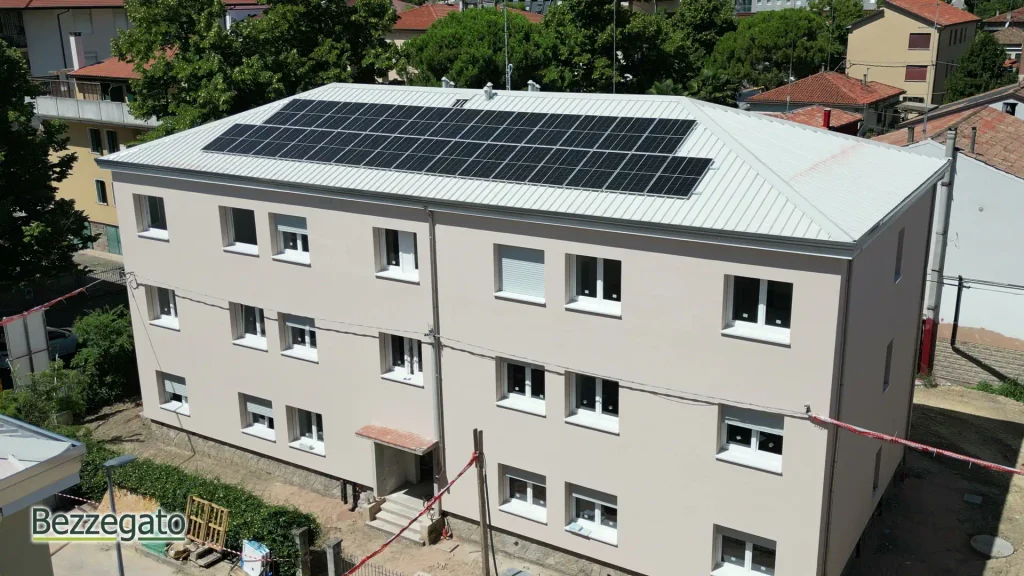 vlcsnap-2026-10-28-18h43m04s557_web | Bezzegato Aerial view of a beige, three-story apartment building with a large solar panel array on its white metal roof, in a green, residential area.