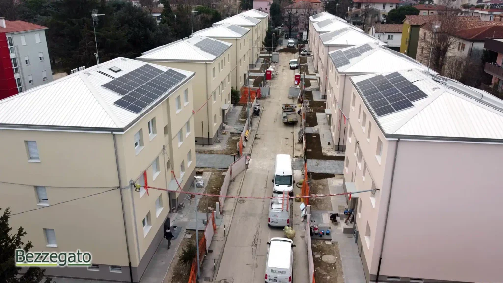 vlcsnap-2026-10-28-18h43m53s172_web | Bezzegato Aerial view of a construction site between new beige apartment buildings with solar panels on the roofs, orange fencing, and white vans along a narrow street.
